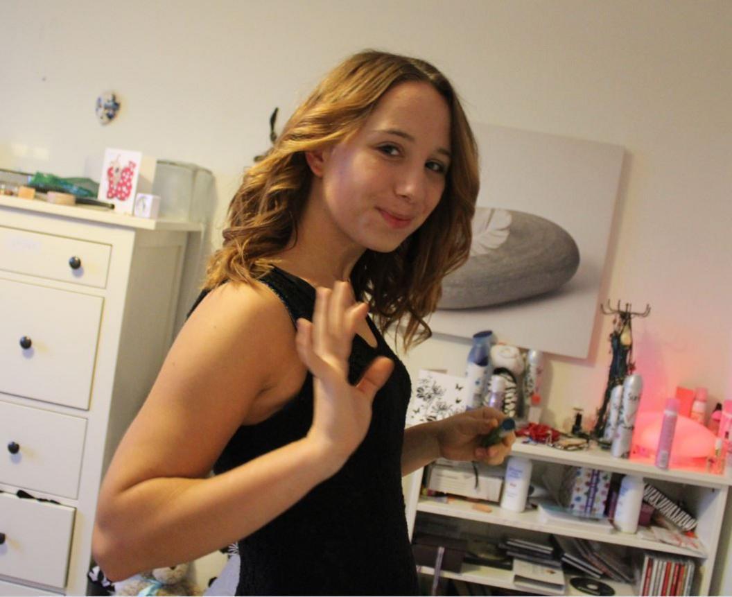 Alice is standing in her bedroom on her 18th birthday. She is wearing a black sleeveless dress and is holding her hand up as if she is waving. There is a shelf and chest of drawers behind her and an artwork on the wall.