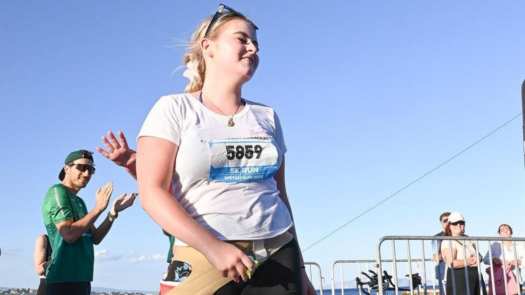 A girl with tied up blonde/brown hair is wearing white top with a race number pinned to it and she is smiling. People behind her are clapping.