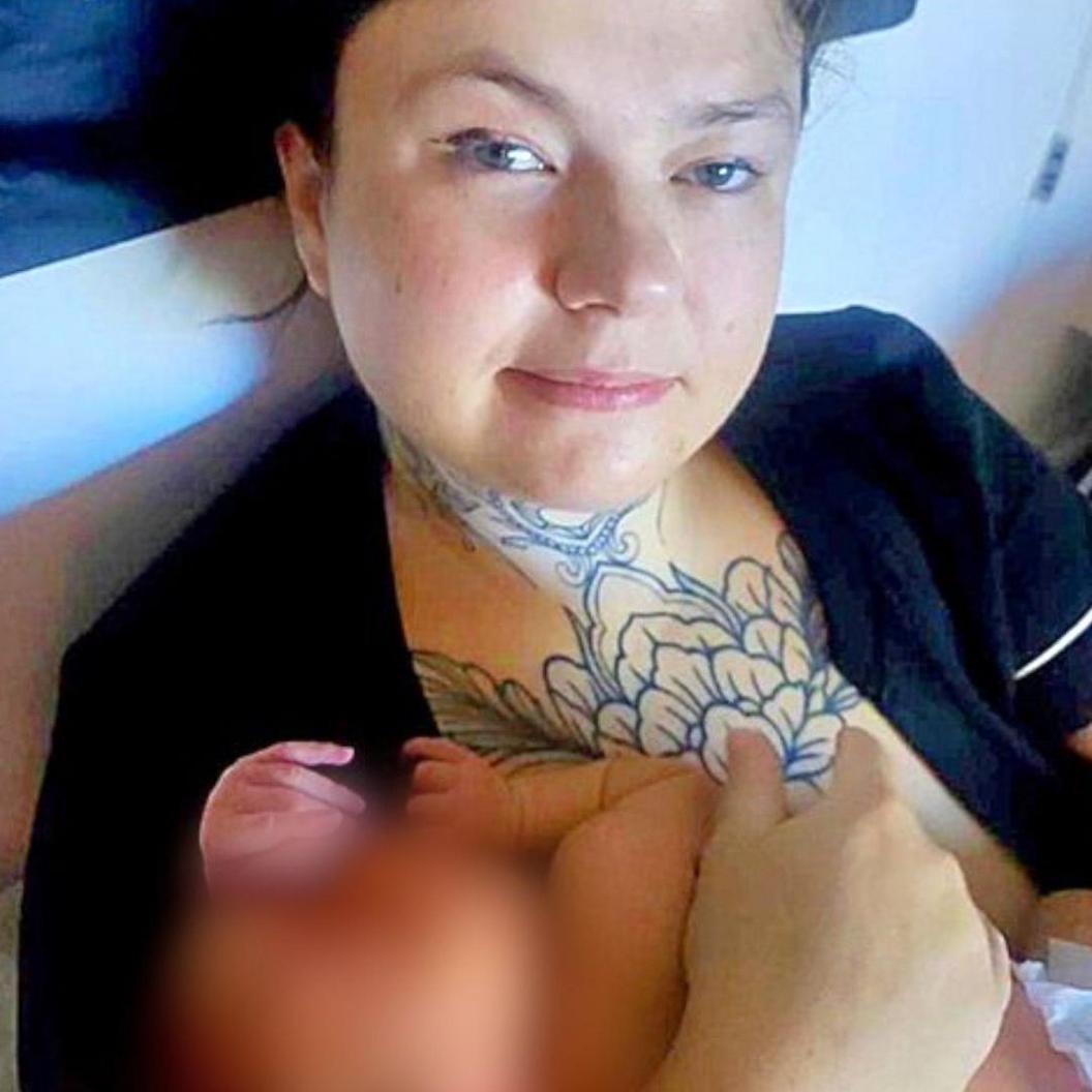 A woman with dark hair holding a baby, looking directly at the camera. She is wearing a black top and has tattoos on her neck and chest.