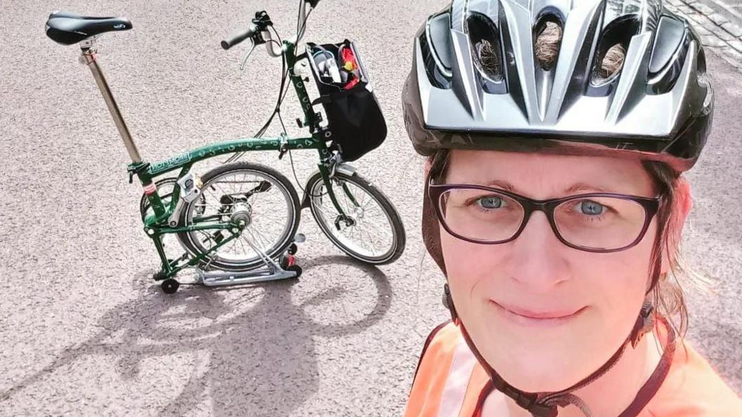 Cyclist Louise Whittle in a selfie. She is wearing glasses, a cycling helmet and an orange hi-vis top. Her green bike, one of those ones that folds up, is on the road behind her.