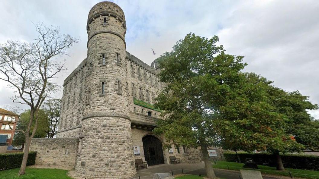 Street view of The Keep Military Museum. It is a large stone gatehouse, resembling a medieval castle with round turrets, crenulations and arrow slit windows.