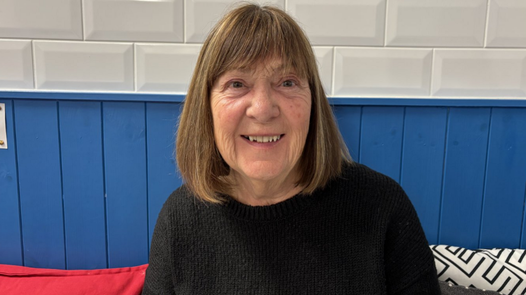 A woman wearing a black jumper smiles a the camera. She is sitting on a bench in a cafe. Blue wooden cladding is behind her. 