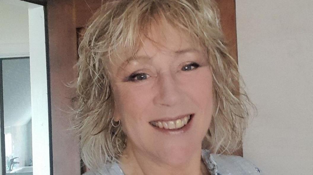 Angie Balwako, a woman with curly blonde hair, is smiling for the camera. She is wearing a light-coloured blouse and small hoop earings.