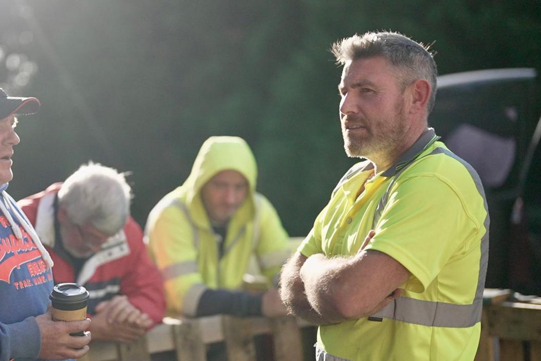 Martin Barry, with short grey hair and beard, arms crossed and wearing a high-visibility jacket, chats to three other protesters leaning on a barricade