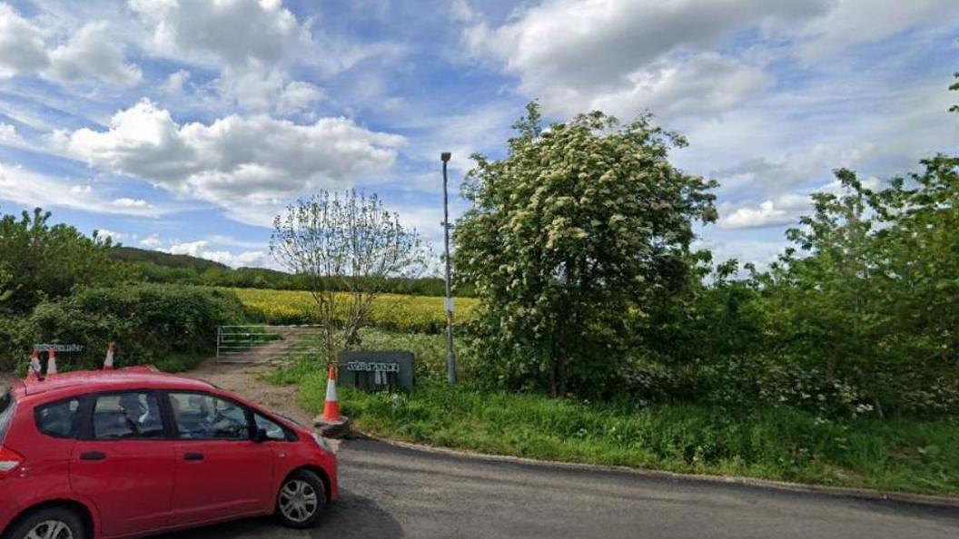 A red car is parked on a road overlooking countryside and a farm gate leading to a field. There are shrubs on the grassy verge and there are traffic cones placed near the gate.