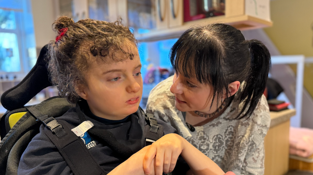 Amelie, who is strapped to a specialist device, has curly hair tied back and is holding her hands together as her mother looks on.