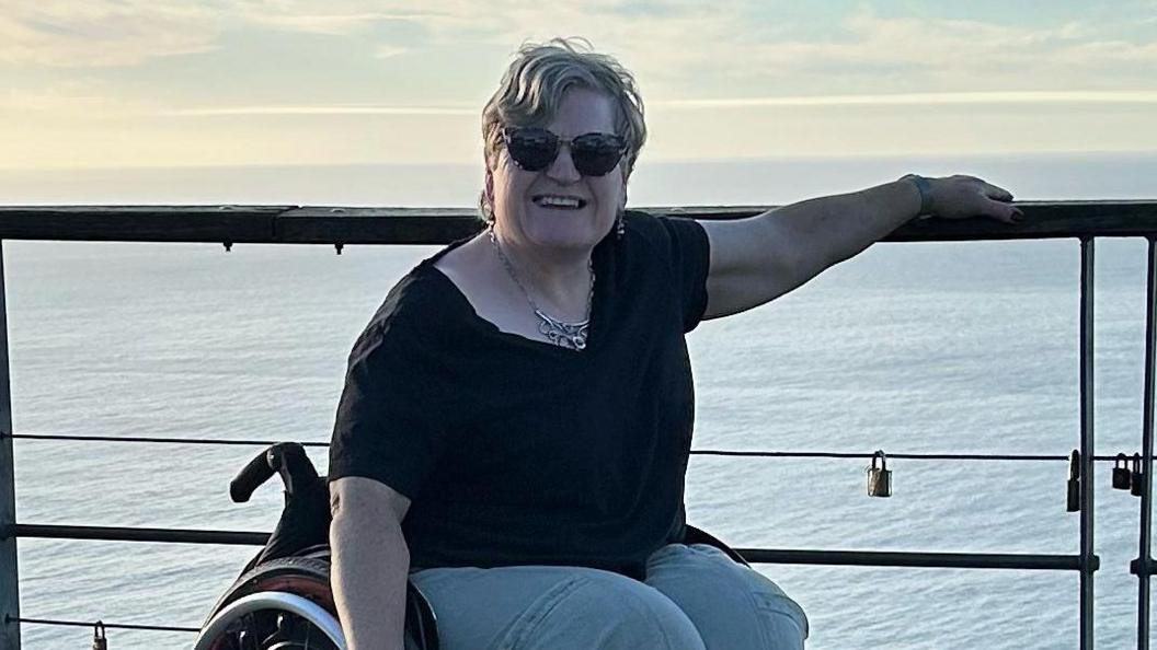 A white woman in a wheelchair smiles and she sits next to a cruise ship's safety fencing