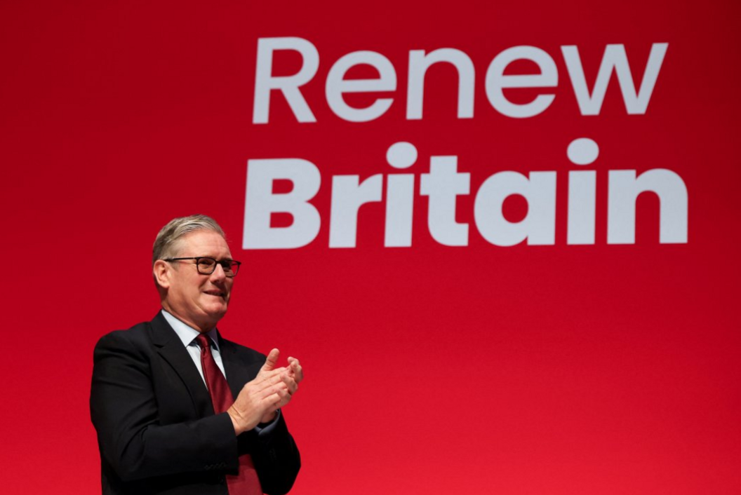 Sir Keir Starmer stands in front of a giant red screen with the slogan Renew Britain. He is wearing a dark suit with a white shirt and a red tie. He is applauding Chancellor Rachel Reeves, not shown.