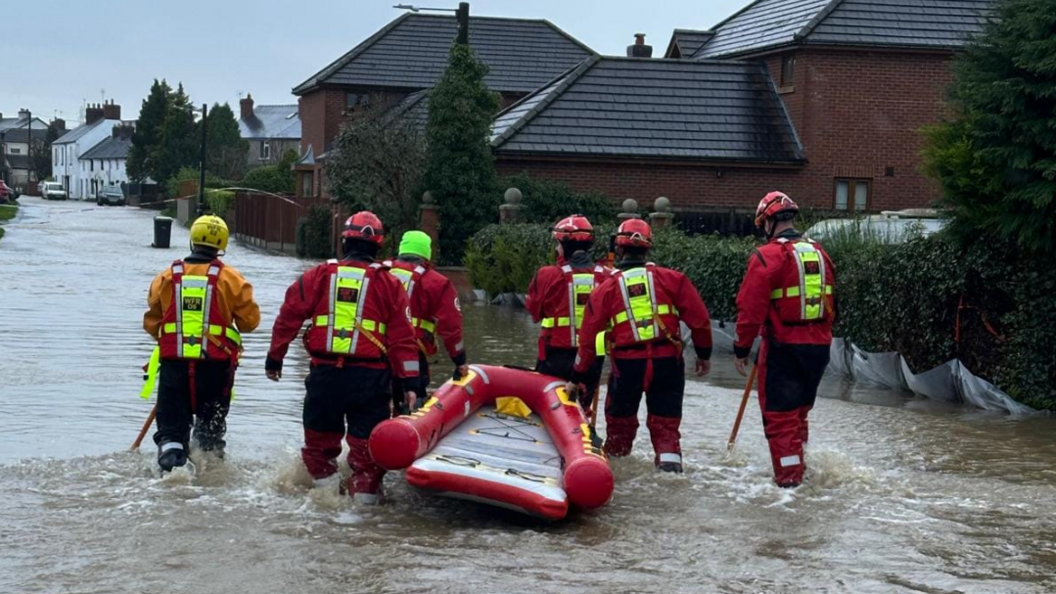 A group of men in red jackets and yellow helmets pulling a red boat through a flooded street with a number of houses in the background