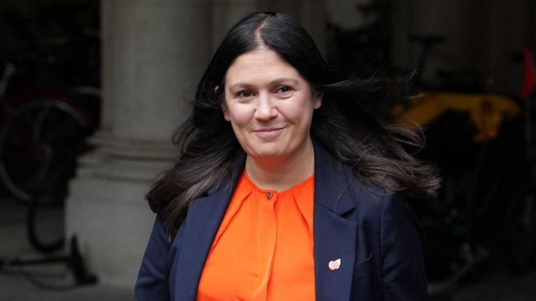 Nandy has long dark hair and is wearing an orange blouse and navy jacket with a poppy pin on the lapel. She is walking outside past a grey stone column.