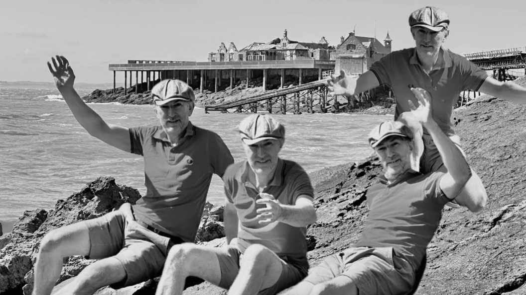 Four men in flat caps raising their hands on the coast with Birnbeck Pier in the background