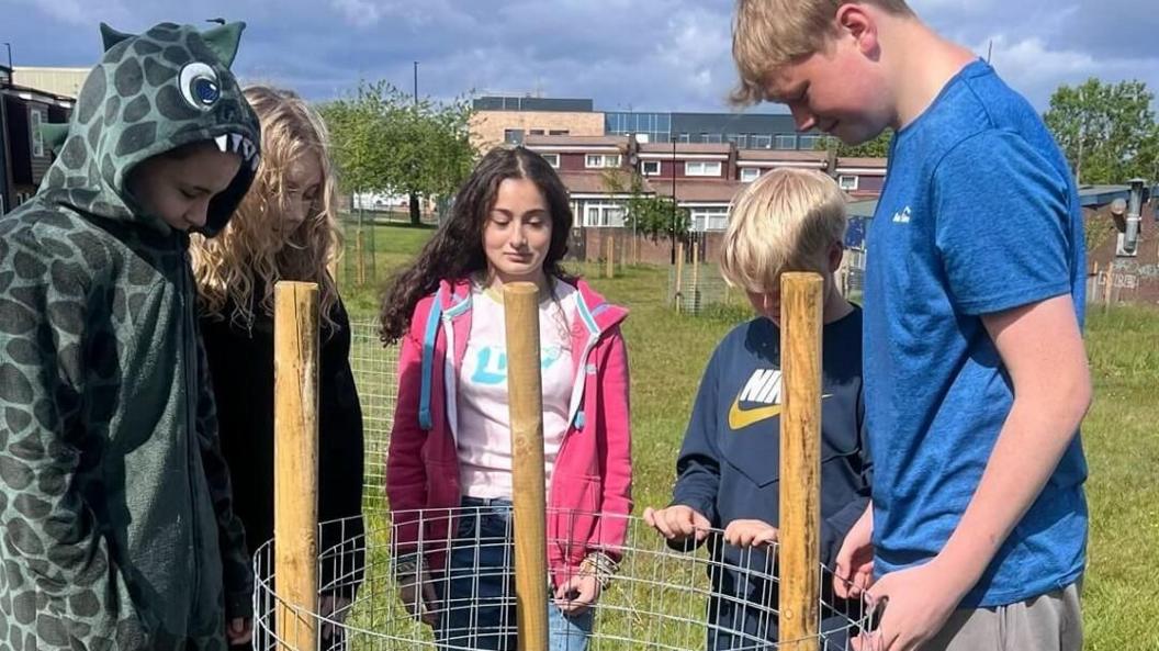 Five youngsters looking at the spot where they had planted a fruit tree. The planting spot has metal mesh and three wooden poles. It is on a field with other similar planting structures dotted around in the distance. There are a number of relatively modern two-storey terraced houses on the edge of the green.