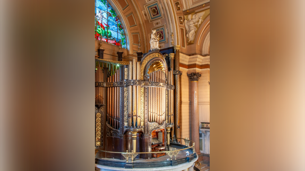 A gold grand organ sits inside a hall with a stained glass window above it. 