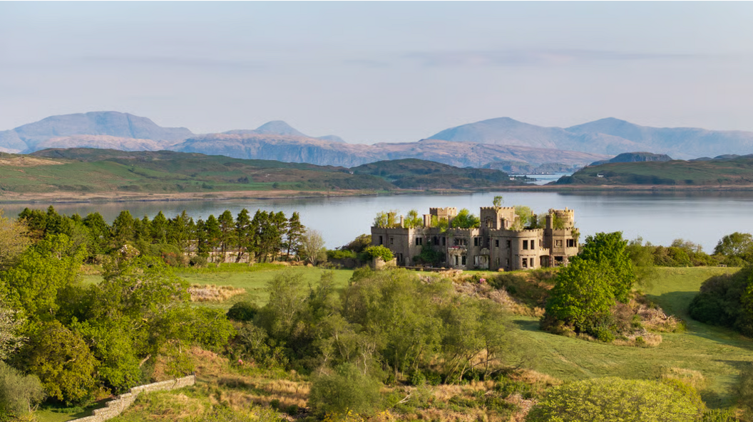 The ruins of Shuna Castle, a flat-roofed turreted castle, are surrounded by grassy banks and trees and overlooking Loch Melfort with a view in the distance of mountains.