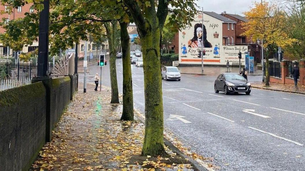 Scene showing a road with cars travelling across. There are autumn leaves on the ground and trees either side of the road. To the right, a mural to King William III.