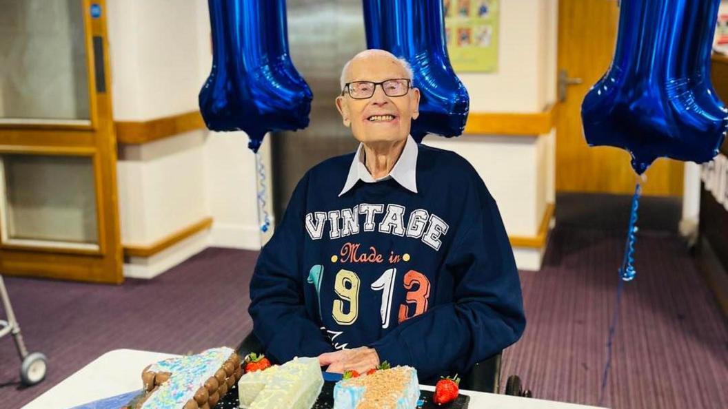 John Farringdon smiling at the camera in a blue 'vintage made in 1913' jumper, with three cakes spelling out '111' in front of him and blue balloons behind him