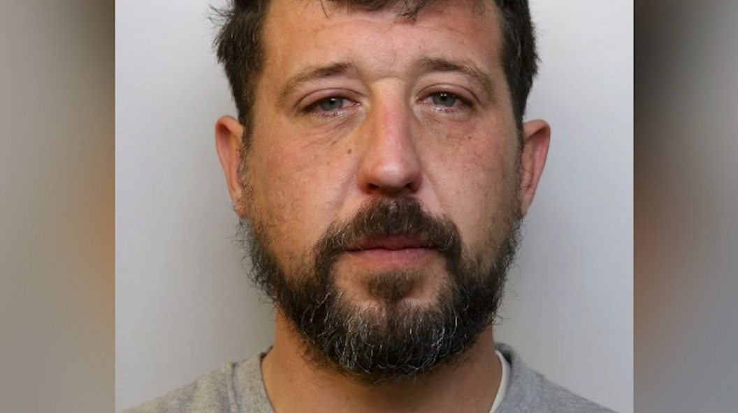Mugshot of Atkinson. He has dark hair and a dark beard and is wearing custody uniform.