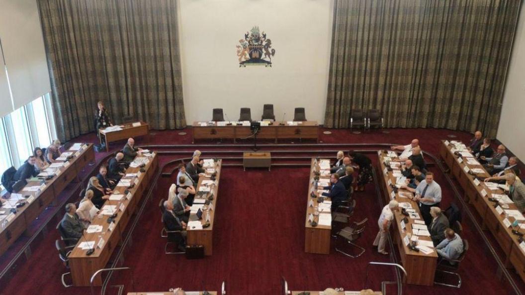 Councillors sitting in the chambers used by Redcar and Cleveland Council for its main gatherings. It has a dark red carpet and wooden benches, with large grey-checked curtains hanging from the windows