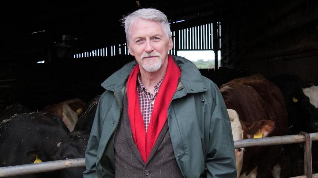 Huw Irranca-Davies, deputy first minister and cabinet secretary for climate change and rural affairs stands on a farm with cows in the background.
