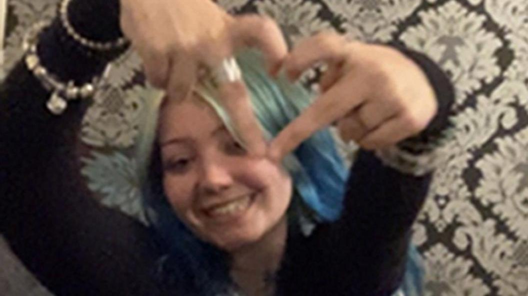 An image of Morgan Dorsett, who has blue hair and is smiling at the camera while holding her fingers in a heart shape up at the camera. 