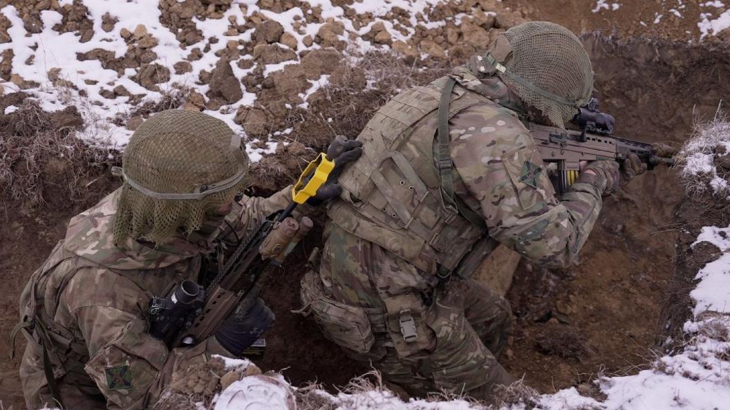 Two soldiers in camouflage army uniforms, holding guns and crouching down in snow-covered trenches 