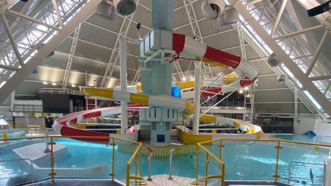 Oasis Beach Pool in Bedford, showing two large slides, one red and one yellow, a pool, with barriers, a large white pyramid roof, and metal frame, around the building.