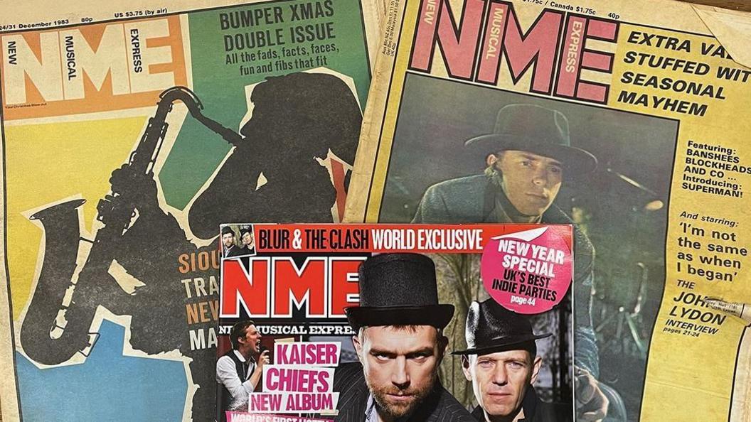 Three NME music magazines overlaying each other on a table. Two at the bottom are from mid-20th century with the paper looking very aged. The third is from the 90s