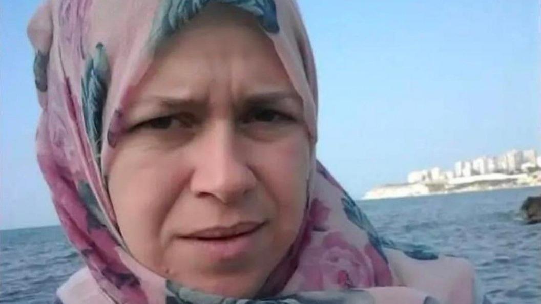 Ghinwa Muhammad Azzam looking into the camera, she is wearing a pink and blue floral headscarf. The sea and coastal buildings are in the background.