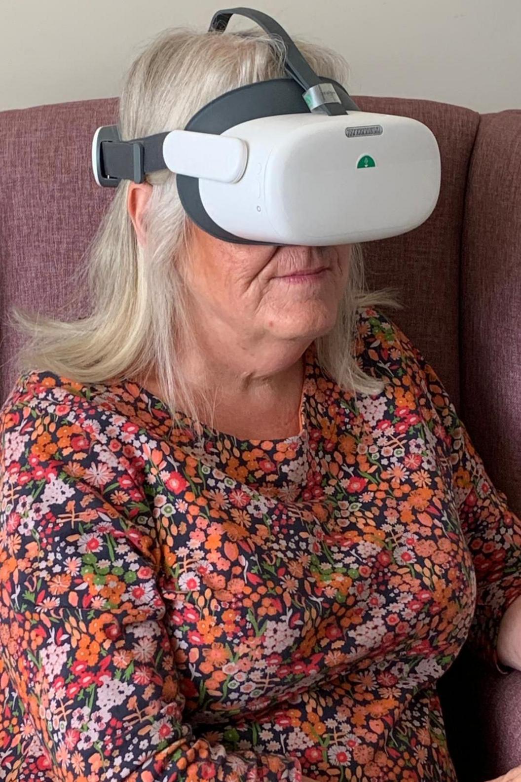 Lady with a patterned dress sat on a maroon sofa and wearing a VR headset.