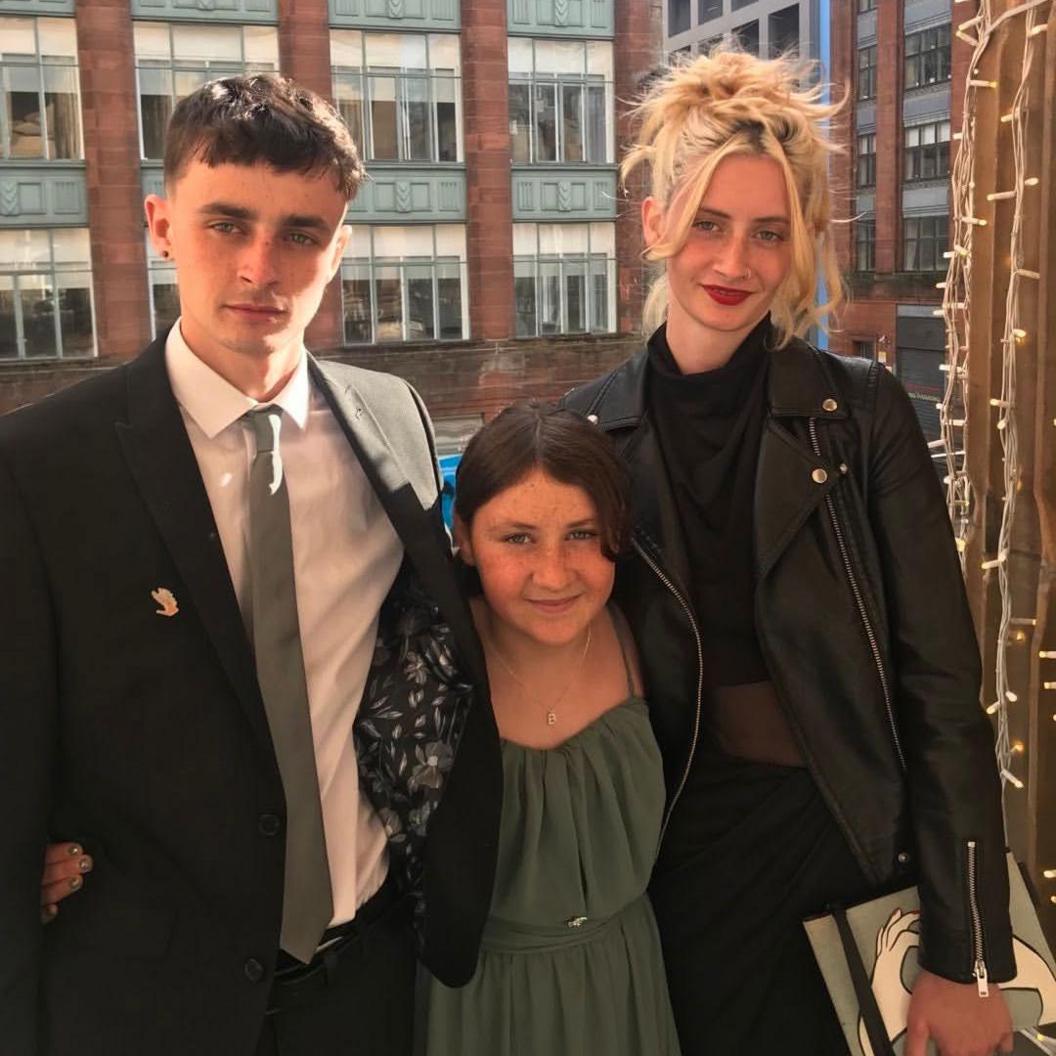 A young man with brown hair in a black suit jacket, a white shirt and grey tie stands next to a small girl with brown hair and a green top, and a young woman with tied-up blonde hair, a black blazer and black jumper.