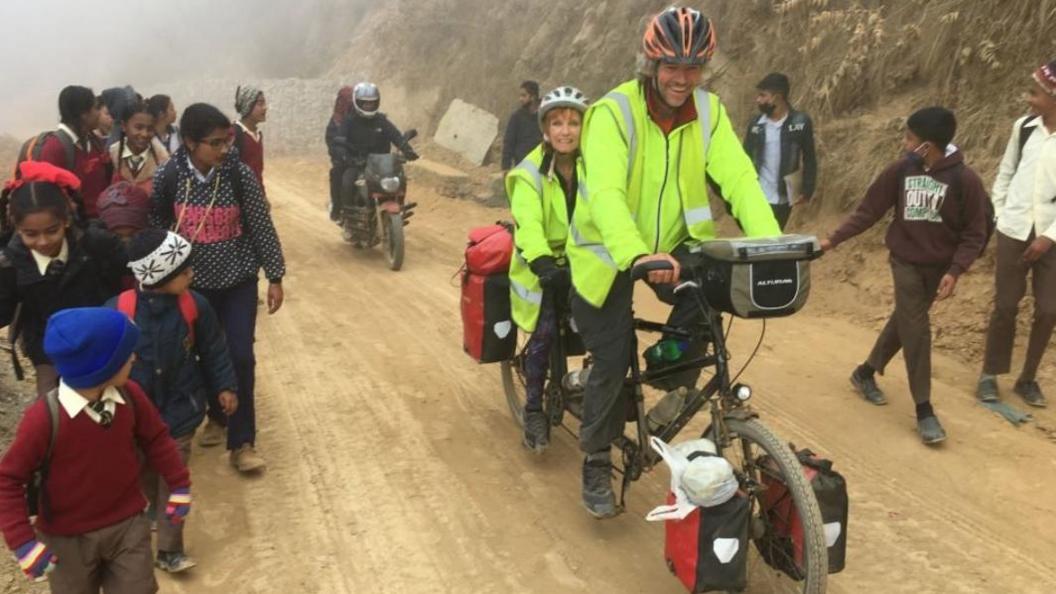 Dave and Helen cycle on their Tandem on a mud road, surrounded by children and a motorbike behind them