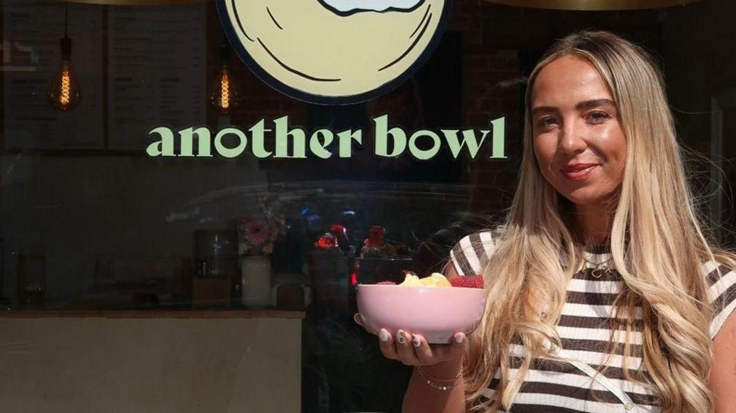 Chloe Marsh stands outside her cafe holding a pink bowl filled with fruit. She has long blonde hair that is lightly curled at the ends. She wears a striped black and white top.