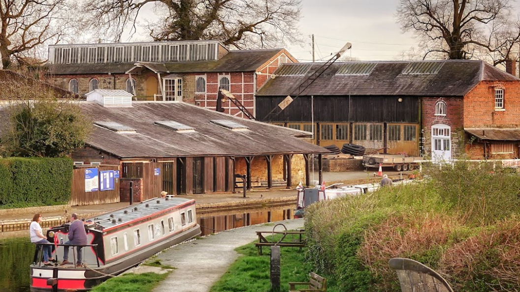 A scenic shot of the yard and its surrounds with two people sat on a barge on the canal in front of buildings at the historic yard