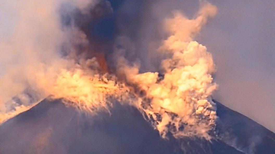 A closer view of Mount Etna during its eruption. Smoke billows from the volcano and some patches of red can be seen within the smoke.