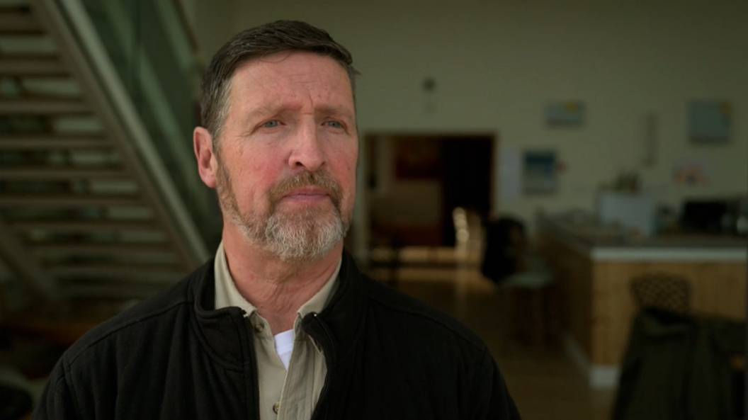 A waist-up portrait shows Michael inside a contemporary building. The individual wears a dark zip-up jacket over a light shirt. Behind him, an open-plan interior includes a staircase, seating areas and wooden furnishings, all softly out of focus to keep attention on the subject.