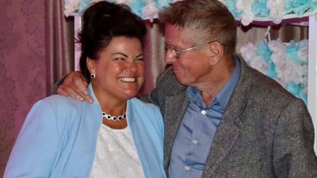 A caucasian woman with dark brown hair and tanned skin wears a white tshirt, blue blazer and sparkly jewellery and grins cheekily at her father, a caucasian man in his 60s or 70s with short brown hair glasses who is wearing a grey blazer and blue shirt.