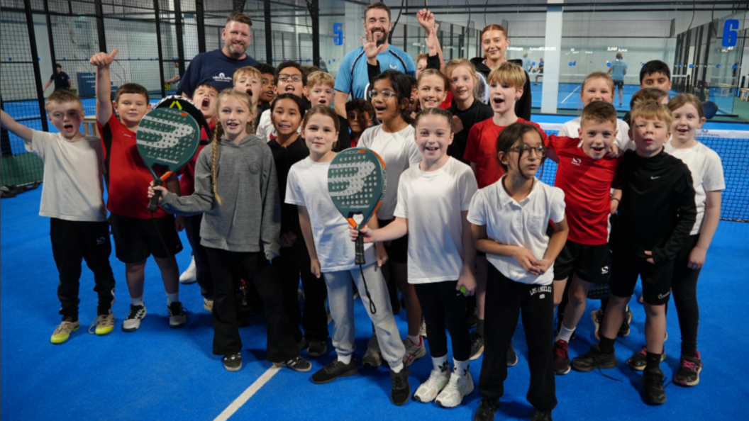 A group of adults and children stand on a racket court, and smile and cheer for the camera.