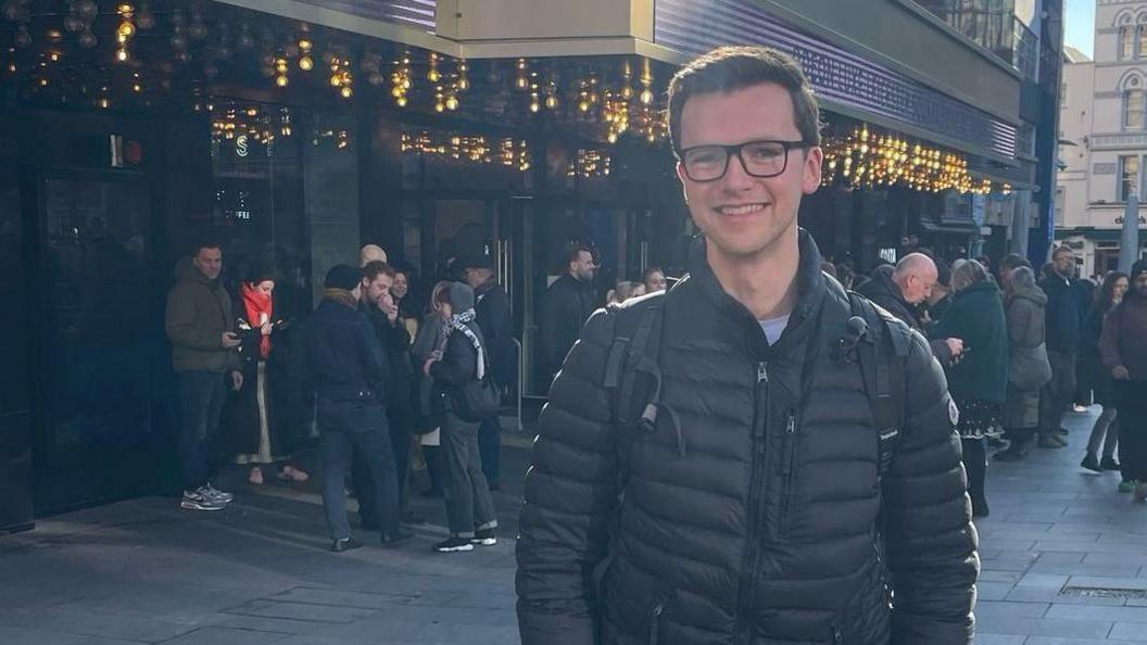 Dan, who has short dark hair and glasses and is wearing a black puffy jacket and black backpack. He is smiling and looking at the camera and behind him is the Odeon cinema in Leicester Square, with twinkling lights and crowds of people outside.