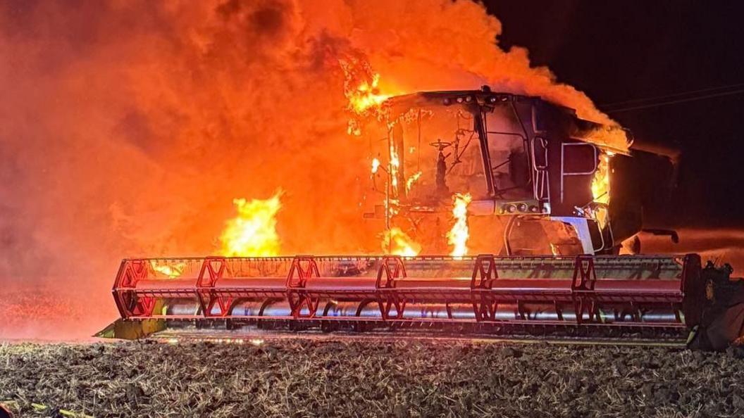 A fully alight combine harvester covered in orange flames which are blowing towards the left. It is night time, the sky is black, and the machine is in a field of stubble.
