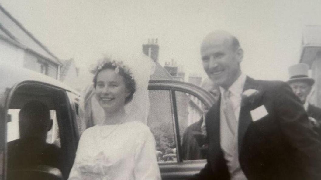 A vintage black and white image of a bride and groom. The bride weas a white dress and a veil, while the groom is in a black suit and waistcoat. They are getting into a car.