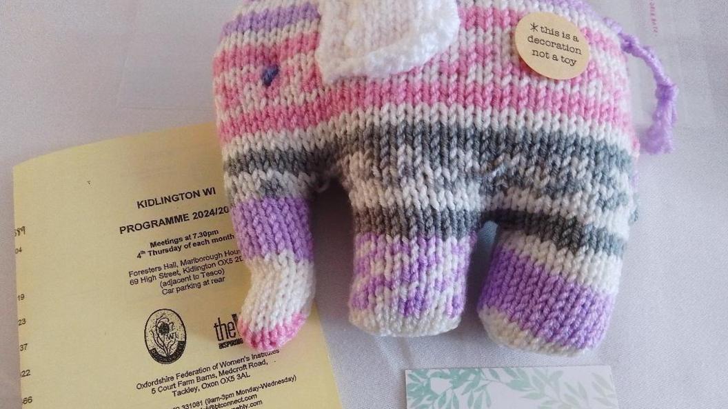 A knitted pink, purple, white and grey elephant resting on a table with a piece of paper with notes on