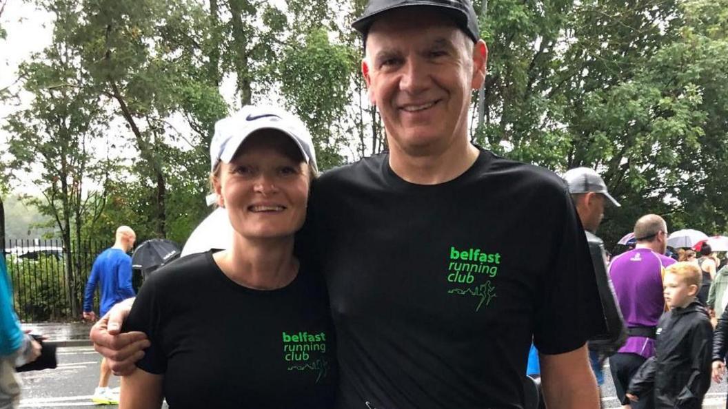 Maria Mulgrew and her husband Peter.  He is standing with his arm around Maria's shoulder and they are both smiling at the camera.   They are both wearing baseball caps and matching black T-shirts with "Belfast Running Club" emblazed in bright green lettering. 