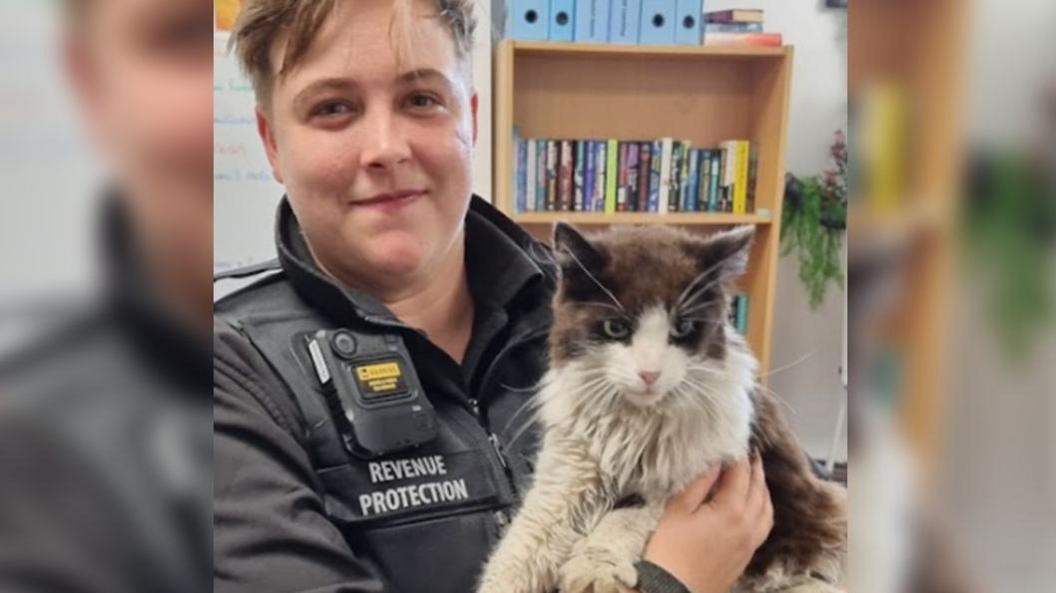 A revenue protection officer wearing a black uniform holding up the black and white cat and smiling