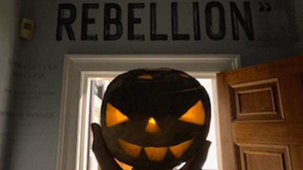 A turnip has been carved into a classic Halloween face, which is glowing from the light of a candle inside. The turnip is being held up framed by a doorway behind, with the word REBELLION in block lettering above the door frame written on the wall.