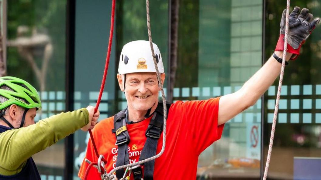 Nigel Haselden, wearing a white safety helmet and red T-shirt, on the ground after his abseil, waving