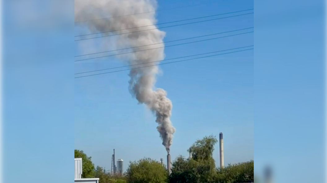 A spout from the oil refinery billows out smoke into the blue sky with trees in the foreground