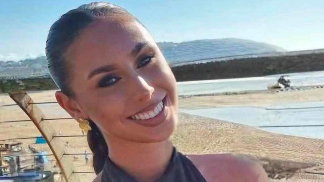 Erin has brown hair tied back and smiles at the camera. There is a beach behind her