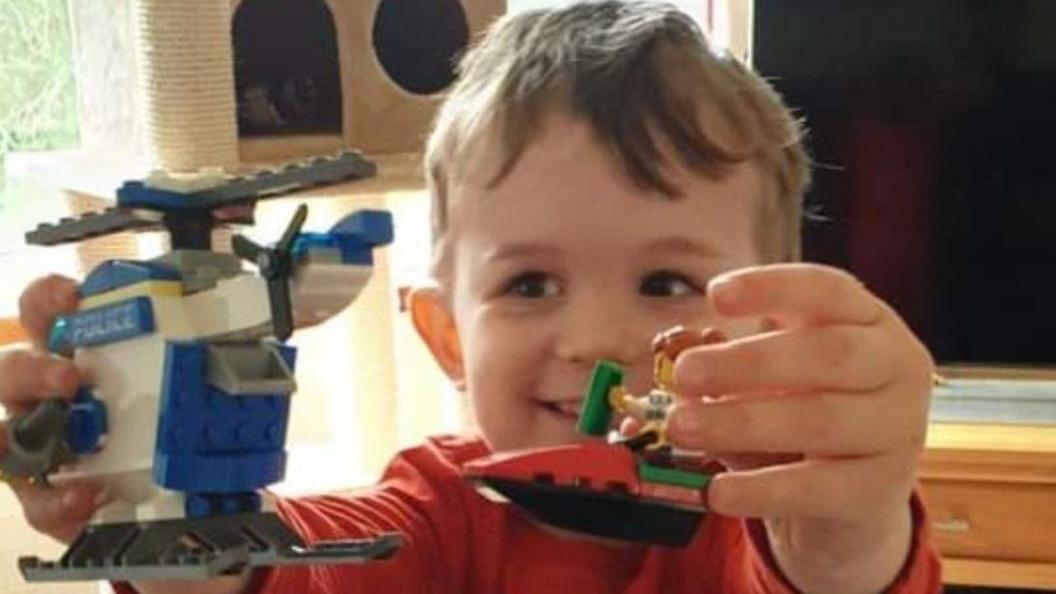 Alexander smiles at two Lego figures: one a police helicopter, the other a figure on a boat. He has brown hair and is wearing a red long sleeved top in a lounge in front of a TV and cat gym. 