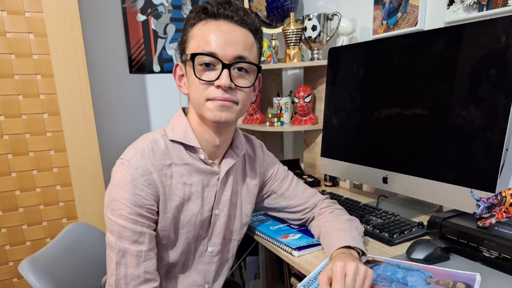 Kenan sitting at a desk. His arm is resting on his own manifesto and he is looking into the camera. There are trophies and a Spiderman ornament on a shelf behind him.