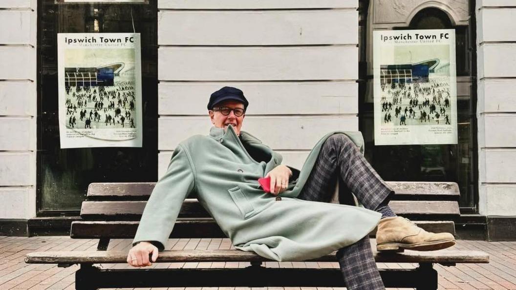 Simon Farr, lying down on a bench, with his foot up, wearing checked trousers and a green coat, between two Ipswich Town posters
